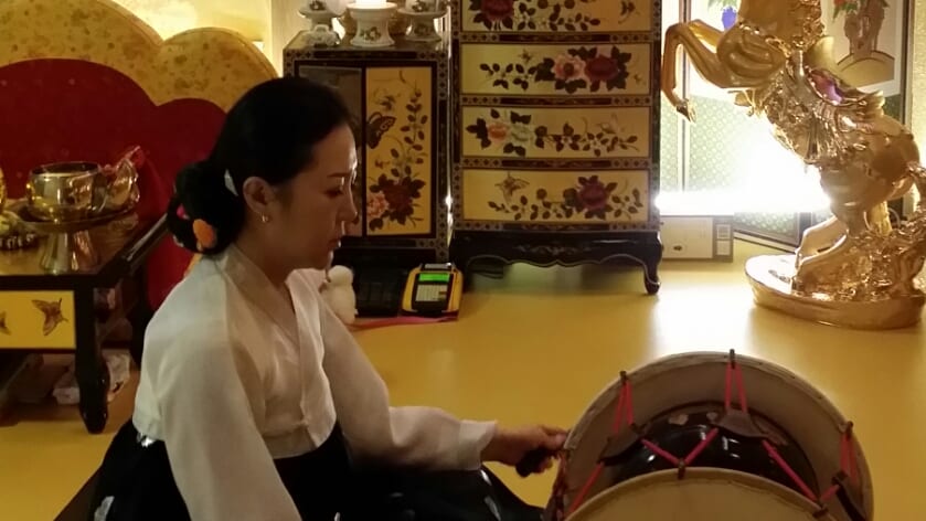 병풍과 자개장이 놓인 신당에서 장구를 치는 당주보살